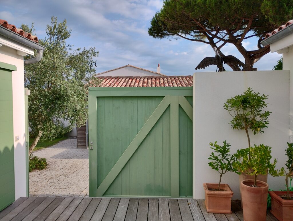 Vue patio et carport
Terrasse en bois
Portail coulissant vert olive entrouvert 
Citronnier 
Cormoran
Pin parasol